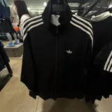 Campera deportiva Adidas Originals de color negro, con cierre frontal, cuello alto y las icónicas tres tiras blancas en los hombros y mangas. Presenta el logo del trébol bordado en blanco en el pecho.