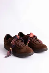 Zapatillas de gamuza color marrón con suela de goma al tono. Presentan cordones planos y un detalle decorativo de pañuelo estampado con cuentas metálicas doradas en la parte frontal.