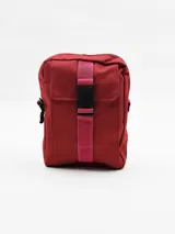 Morral color bordó con cierre superior y bolsillo frontal con solapa, asegurado con una tira de tela rosa y hebilla plástica negra.