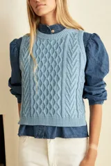 Chaleco de punto celeste claro, sin mangas y con cuello redondo. Presenta un diseño de punto trenzado (ochos o aranés) en el centro de la delantera y un borde acanalado en el ruedo.