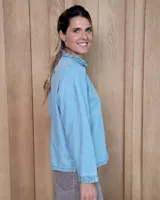 Camisa de denim con cuello alto tipo mao con volados, cierre frontal con botones y mangas largas con puños elásticos con volados.