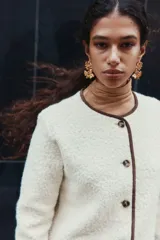 Saco corto de tejido sherpa color crema, con cuello redondo y cierre frontal de botones marrones. Presenta ribetes de tela marrón en el escote y la tapeta frontal.