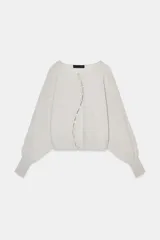 Chaqueta de punto color beige claro, de cuello redondo y manga larga abullonada con puño ajustado. Cierre frontal cruzado con botones plateados.