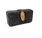 Clutch rectangular de estructura rígida, confeccionado en tejido de textura rústica color negro con efecto gastado. Presenta un aplique central en forma de piedra ágata en tono tostado con borde dorado y cierre magnético. Incluye una cadena metálica dorada desmontable.