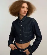 Camisa de jean estilo cropped de color azul oscuro, con cuello inglés, mangas largas, cierre frontal de botones metálicos y un bolsillo aplicado en el pecho. Cuenta con costuras pespunteadas en tono contrastante.