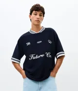Remera de tejido texturizado color azul marino, con cuello redondo y mangas cortas con puños a rayas blancas. Presenta bordados frontales con la inscripción 'Future Co.', el número '98', un logo ovalado y estrellas.