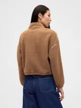 Buzo de polar tipo sherpa color marrón, con cuello alto y cierre frontal parcial. Presenta el logo de la marca bordado en el pecho y detalles de costuras reforzadas en hombros y mangas.