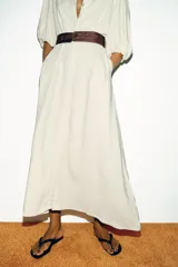 Vestido midi color crudo, con cuello camisero, mangas abullonadas por debajo del codo con puños elásticos y cierre frontal oculto con botones. Incluye bolsillos laterales y un cinturón ancho de cuero marrón oscuro con hebilla metálica.