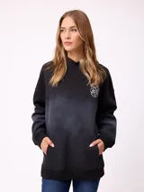 Canguro oversize color negro con efecto degradado, capucha, bolsillo frontal tipo canguro y puños elásticos. Presenta un parche con logo en la manga.