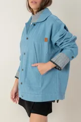 Campera de jean oversize color azul claro, con cuello camisero, cierre frontal con botones y bolsillos frontales. Presenta un diseño de corte amplio y cómodo con mangas largas.
