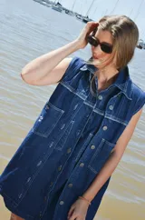 Vestido corto de jean color gris oscuro con efecto acid wash, sin mangas, con cuello camisero y cierre frontal de botones. Incluye dos bolsillos en el pecho con solapa y pespuntes decorativos.