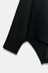 Jersey negro de tejido punto, con cuello redondo y corte tipo capa asimétrico, más largo en la espalda.