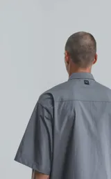 Camisa de manga corta de poplin, color gris, con silueta oversize. Presenta dos bolsillos delanteros, uno con solapa y otro con ojal y botón.