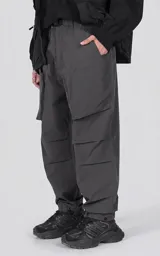 Pantalón cargo oversize de color gris, confeccionado en tejido técnico ripstop. Cuenta con cintura elástica con presillas, cierre frontal con broche técnico, múltiples bolsillos amplios y sistema de ajuste con broches snap en los tobillos.
