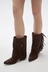 Bota corta de gamuza color chocolate, con punta fina y taco bajo. Presenta detalle de flecos en el lateral exterior y dos correas decorativas con hebillas metálicas en el tobillo.