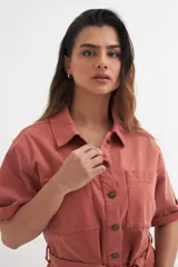 Mono corto color beige con cuello camisero, mangas cortas con dobladillo, cierre frontal con botones, cinturón de tela y bolsillos delanteros.