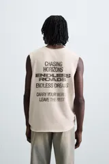 Camiseta tank de punto color beige claro, corte relajado (relaxed fit) y cuello redondo. Presenta un estampado central de un vehículo todoterreno cargado de equipaje conduciendo por una carretera desértica. Los bordes del cuello y las sisas tienen un acabado deshilachado.