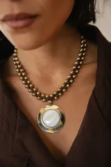 Collar corto con dije circular de nácar engarzado en metal dorado. La cadena está compuesta por una tira de gamuza marrón trenzada con pequeñas cuentas metálicas doradas.