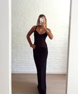 Vestido largo negro, ajustado al cuerpo, con escote en V y tirantes finos. La parte superior del corpiño es de tejido con brillo o lentejuelas, y el resto del vestido es de tela semitransparente o gasa, con un forro opaco que cubre desde la cadera hacia abajo.