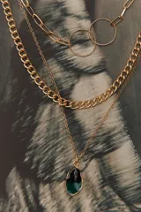 Colgante dorado de tres cadenas de diferentes grosores y diseños. La cadena más larga tiene dos dijes circulares unidos. La cadena más corta tiene un dije de piedra facetada color verde oscuro.