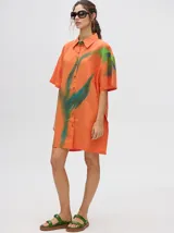 Camisa tipo vestido de corte oversize, confeccionada en viscosa. Presenta un diseño estampado con trazos abstractos en tonos verdes sobre una base naranja, cuello clásico, cierre frontal con botones y mangas cortas.