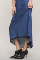 Vestido maxi azul de satén con diseño levemente asimétrico, corte al bies y escote en V con detalle de puntilla negra. Presenta espalda deportiva con tirantes finos regulables y ribete de encaje en el ruedo.