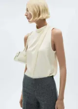 Blusa color crema de tejido fluido satinado, con cuello alto y diseño drapeado sin mangas. Cierre de botón en la parte posterior.