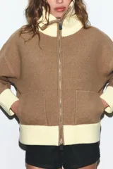 Chaqueta de punto con cierre frontal de cremallera, cuello alto, manga larga y bolsillos delanteros de plastrón. Presenta un diseño bicolor en tonos marrón y amarillo claro en cuello, puños y ruedo.
