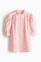 Blusa color rosado con cuello mao y botones ocultos en la parte delantera. Tiene canesú de doble capa con frunces adelante y atrás, mangas cortas abullonadas y puños con abertura y botón.