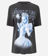 Remera de algodón color gris oscuro (efecto desteñido/vintage) con estampa frontal inspirada en el personaje Glinda, el Hada Buena, de la película Wicked. La estampa incluye el texto "SPARKLE LIKE GLINDA WICKED" y una ilustración de Glinda con su varita mágica.