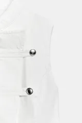Top de denim blanco sin mangas, con cuello mao y cierre frontal cruzado con trabillas y botones metálicos.