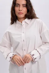 Camisa blanca de corte oversize, con mangas largas y puños anchos decorados con bordados en tono oscuro. Cuenta con cuello camisero clásico y cierre frontal de botones.