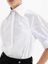 Camisa de popelina blanca con bordados de pequeñas bocas que forman un patrón de rayas. Presenta un diseño de corte holgado, mangas cortas con volumen, cuello clásico con broche metálico y cartera oculta con botones al tono.