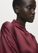 Vestido midi de tejido satinado en color bordó, con cuello alto, manga larga y diseño recto. Presenta una abertura frontal en el bajo y un cinturón ajustable con detalle metálico en la cintura.