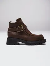 Bota corta tipo borcego de cuero negro con textura granulada, suela track dentada y tira decorativa con hebilla metálica en el tobillo.