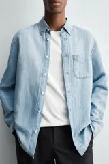Camisa de corte regular fit confeccionada en denim ligero de algodón con efecto lavado. Presenta cuello solapa con botones, manga larga con puños abotonados, cierre frontal de botones y un bolsillo plastrón en el pecho.