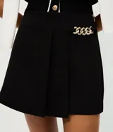 Short pollera de sastrería marca Cortelle, color bordó, confeccionado en crepé. Presenta sobreposición frontal efecto pollera con pliegues y detalle de cadena metálica decorativa en el lateral.