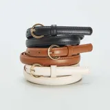 Pack de tres cinturones finos de cuero sintético en colores negro, marrón y blanco, con hebilla dorada en forma de semicírculo.