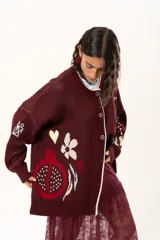 Saco de punto color vino con diseño holgado, cuello redondo y mangas largas. Presenta un gran bordado central en la espalda con forma de flor en tonos crema y rojo, acompañado de un pequeño bordado de sol y una frase bordada en la parte inferior.