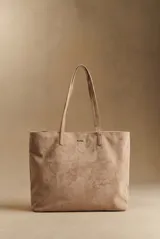 Bolso tipo tote de gran tamaño en color camel, confeccionado en cuero vegano con acabado efecto gamuza. Presenta doble asa larga para llevar al hombro y una placa metálica pequeña con el logo de la marca en el frente.