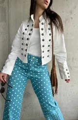 Chaqueta corta de estilo militar en color blanco, con múltiples filas de botones metálicos grabados y detalles de cordones en el frente. Cuenta con cuello mao y mangas largas con botones en los puños.