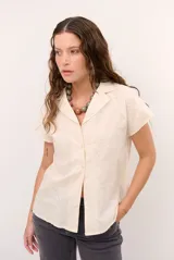 Camisa de manga corta color beige con cuello solapa, cierre frontal de botones y delicados bordados blancos en el frente.