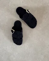 Zueco tipo sandalia de estilo jaula, confeccionado en gamuza negra con tiras entrelazadas y hebilla metálica dorada lateral. Cuenta con plantilla acolchada con logo de la marca.