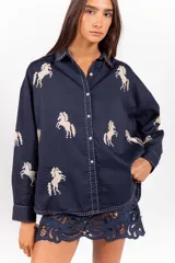 Camisa de corte clásico y calce amplio, color marfil, con cuello camisero, mangas largas y cierre frontal con botones. Presenta bordados coloridos de caballos en contraste distribuidos en la prenda.