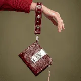 Billetera color bordó con textura símil cocodrilo y detalles plateados. Tiene correa de mano removible, cierre principal, compartimento para billetes, bolsillo con cierre para monedas y ranuras para tarjetas.