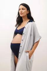 Vestido camisero maternal de color gris claro, con cuello clásico, cierre frontal de botones oscuros, mangas cortas y bolsillos laterales. Presenta un pequeño bordado de corazón en el pecho.
