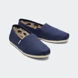 Alpargatas de lona de algodón color crema, estilo slip-on, con elástico en forma de V en el empeine y suela de goma. Presentan la etiqueta de la marca Toms en el lateral.