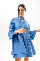 Vestido corto azul, estilo camisero, con cuello camisero, cierre frontal con botones y mangas acampanadas con volados. Presenta un diseño holgado con cortes fruncidos en el cuerpo.