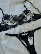 Conjunto de lencería de dos piezas en color negro, compuesto por un soutien con aro y copas de tul bordado con motivos florales, y una bombacha tipo colaless con tiras elásticas y detalle de aplique con letras de strass.