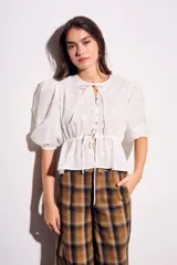 Blusa negra de algodón con diseño de broderie, mangas tres cuartos abullonadas, cierre frontal con botones y detalle de cordón ajustable en la cintura.